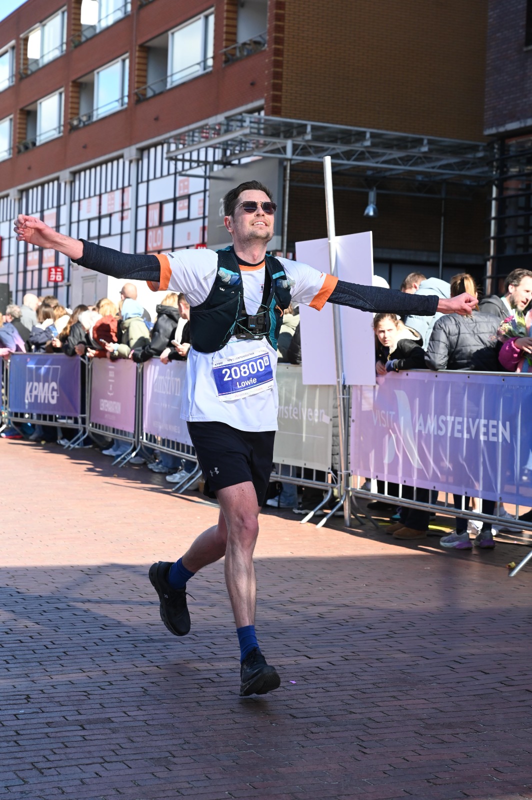 Lowie van Eck runs down a brick street toward the finish of the 2025 KPMG Lentemarathon in Amstelveen, smiling with both arms stretched wide. He wears sunglasses, a white running shirt with orange trim, a dark hydration vest with a small camera on the chest, black shorts and bib number 20800 with his name, Lowie. Spectators line the metal barriers beside him, which carry KPMG and Visit Amstelveen banners, in front of a red brick building.