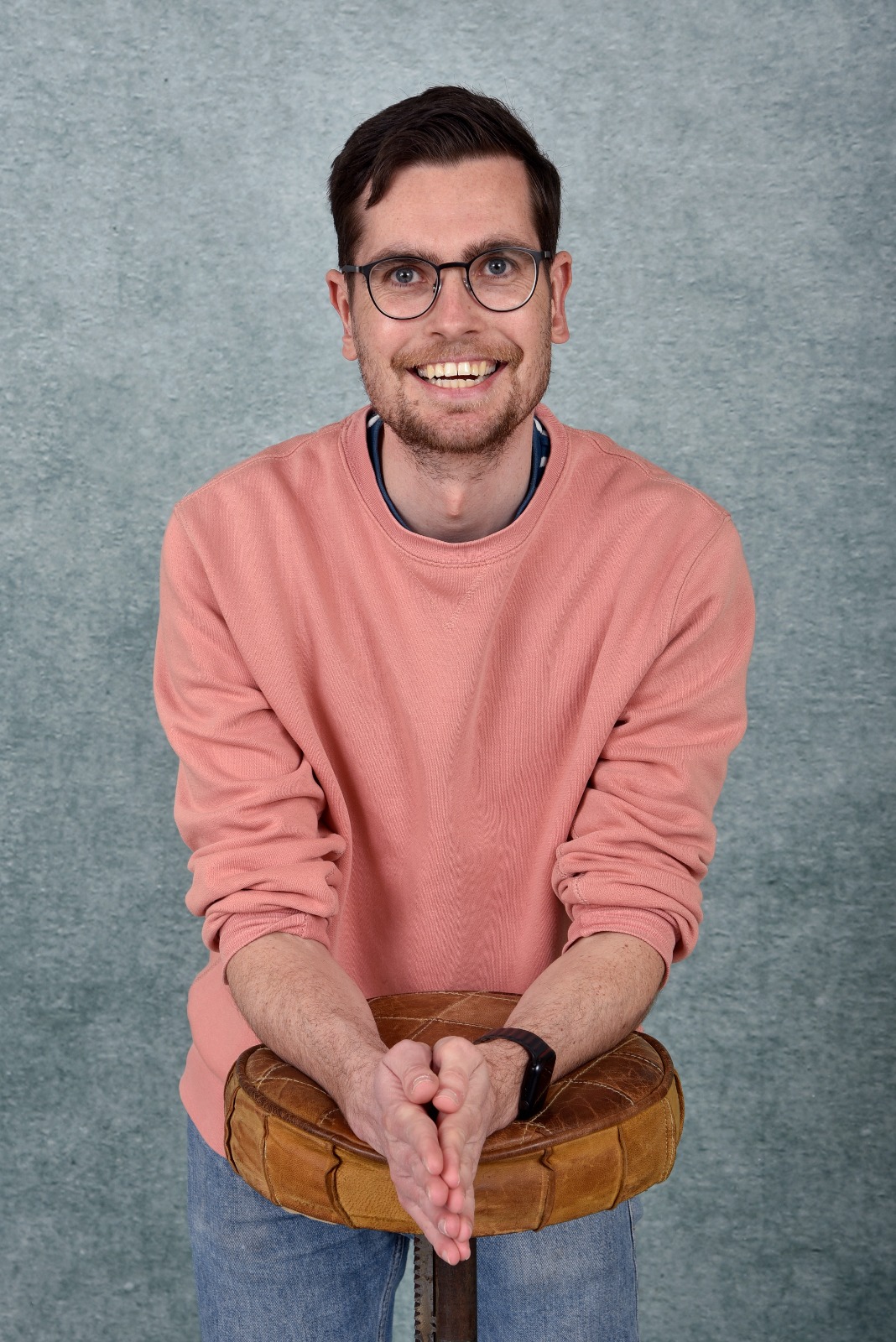 Portrait of Lowie van Eck, smiling widely at the camera. He has short dark hair, a light beard and round dark-rimmed glasses, and wears a salmon-pink sweatshirt over a collared shirt with blue jeans. He leans forward with his forearms resting on a round tan leather stool, hands pressed together, in front of a mottled grey-blue studio backdrop.
